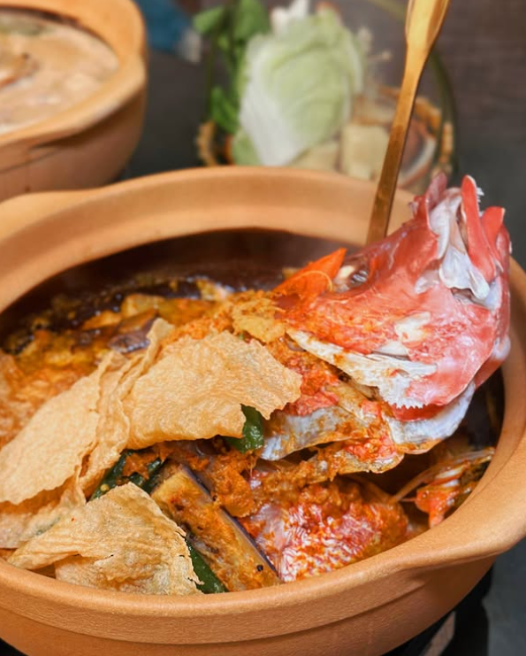 Curry Fish head dish in a ceramic pot with a spoon, surrounded by green vegetables in Clarke Quay Restaurant.