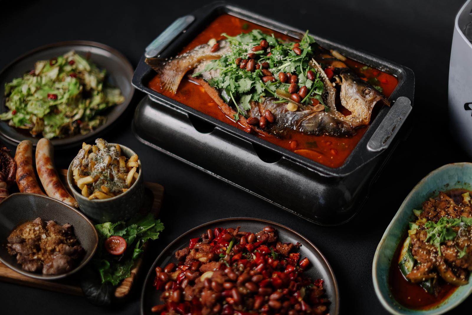 Assorted dishes including a roasted fish dish on a grill, surrounded by various side dishes on a dark surface.