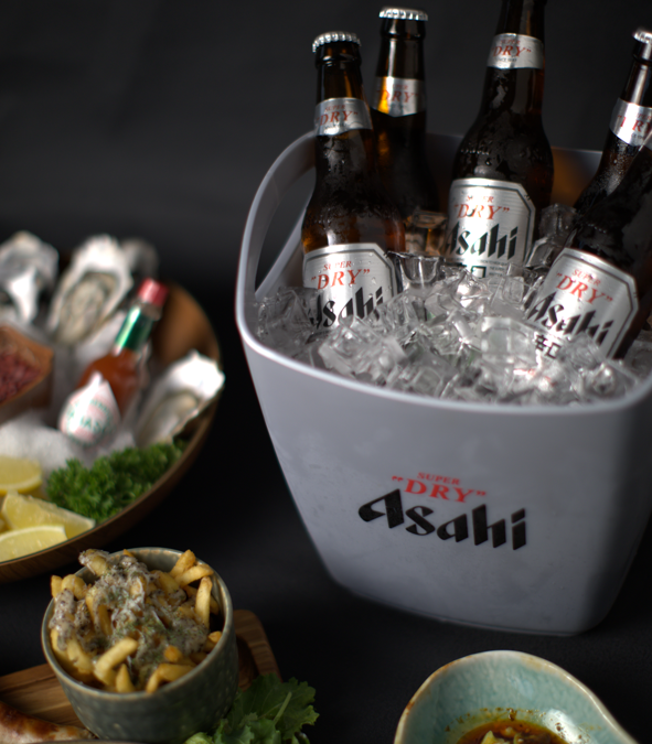 Bottles of Asahi beer in a bucket with ice, surrounded by snacks on a dark background