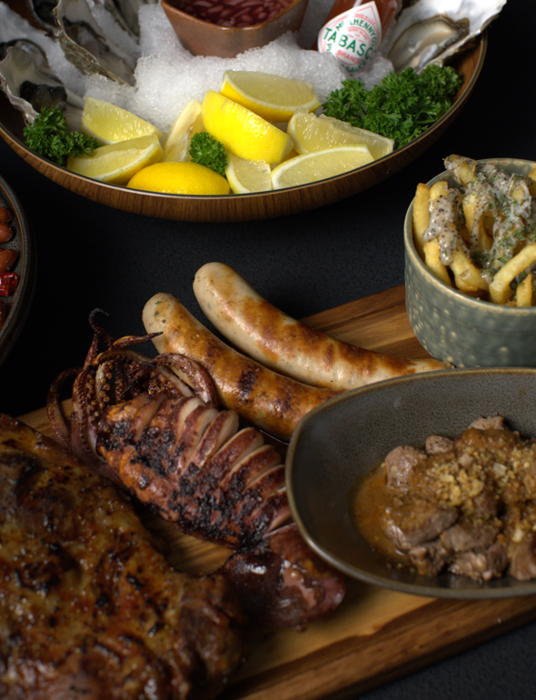 Assorted grilled meats and sides on a dark surface with lemons and a bowl of pasta.