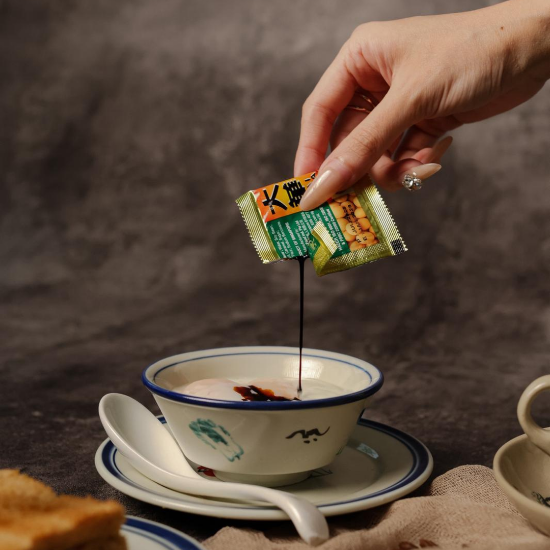 Hand pouring dark sauce from a packet into a  soft boiled egg bowl in  nanyang restaurant.