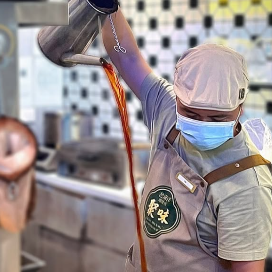 Person in a kitchen pouring a kopi tarik from a faucet, wearing a mask and apron with JUwei logo.
